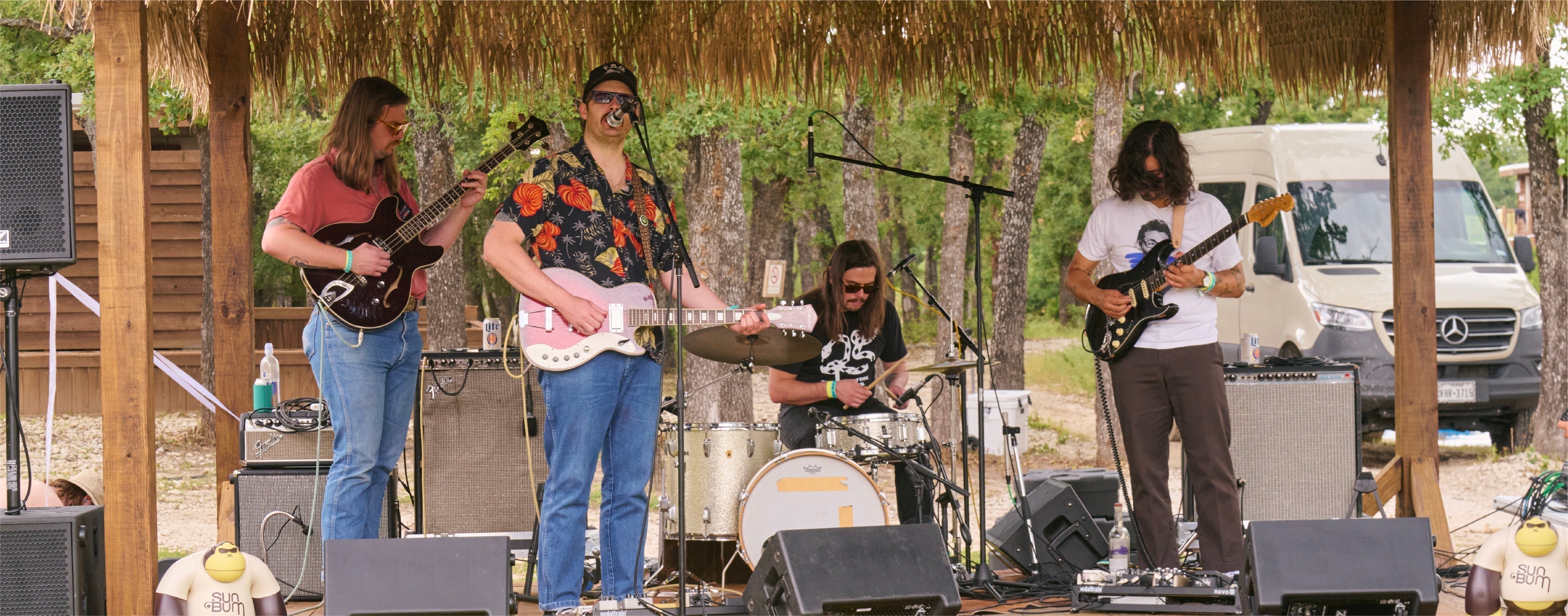 Band performing on a stage with a thatched roof in a natural setting