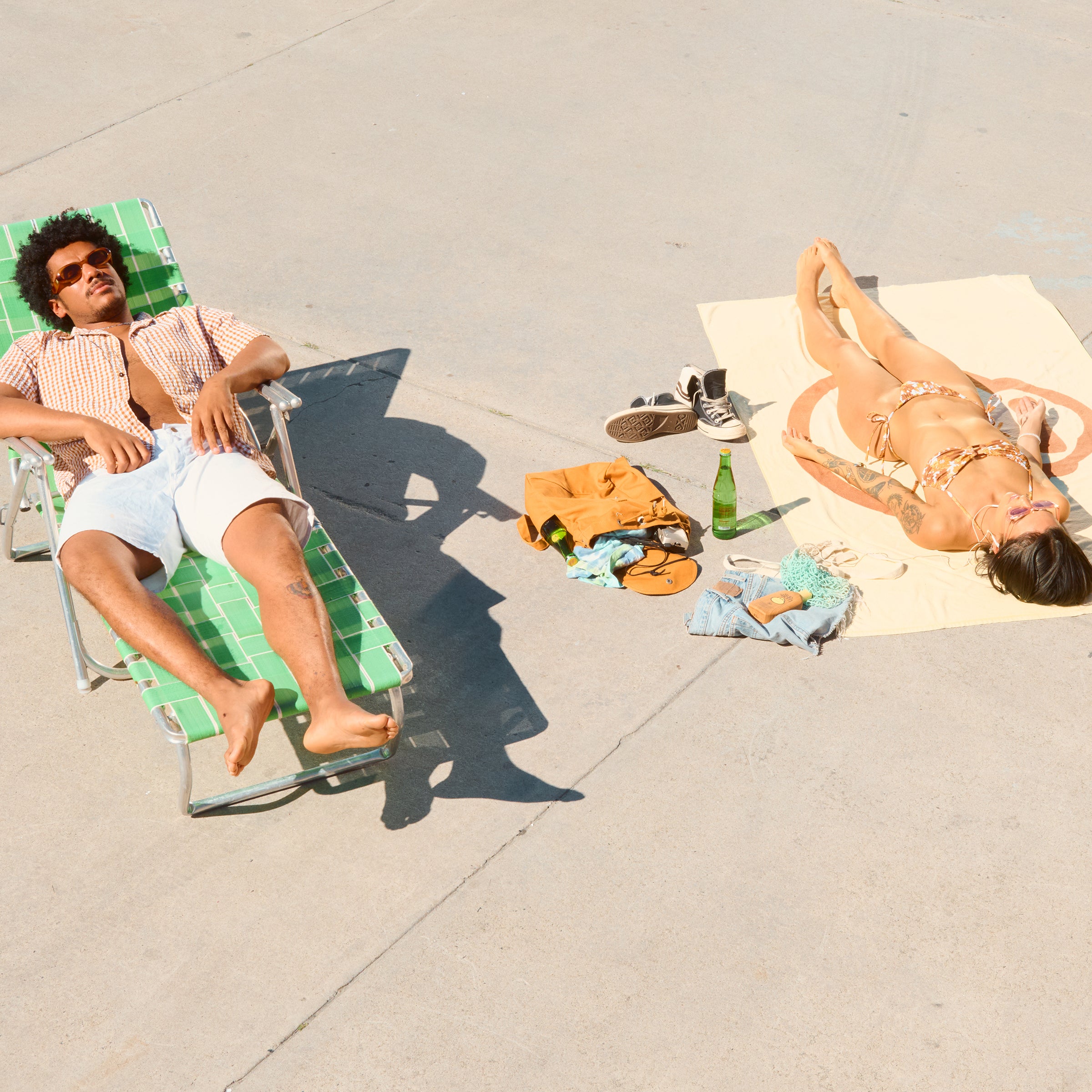 Two people sunbathe on pavement using beach towel and chair, enjoying warm weather vibes – Tanning Collection - Sun Bum
