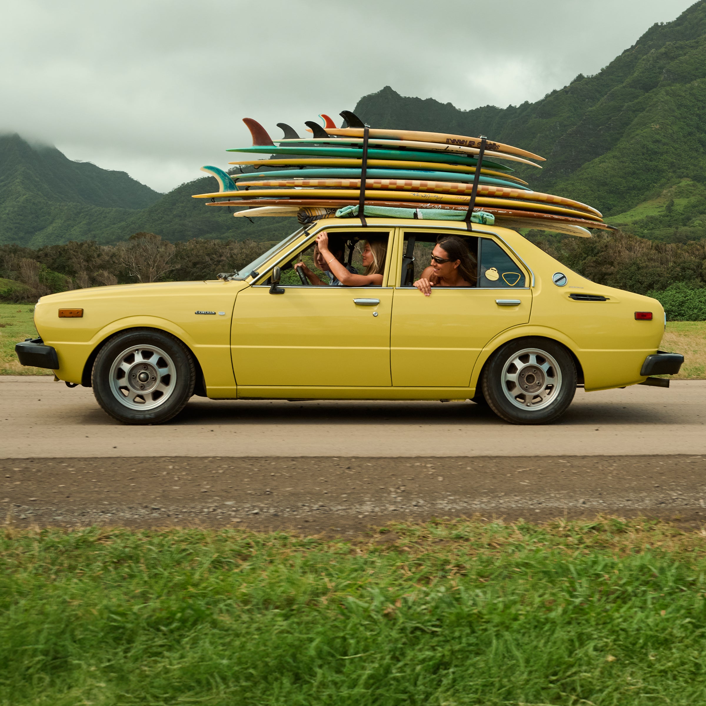 Vintage yellow car loaded with surfboards cruises past green mountains on a tropical road trip – Tanning Collection - Sun Bum