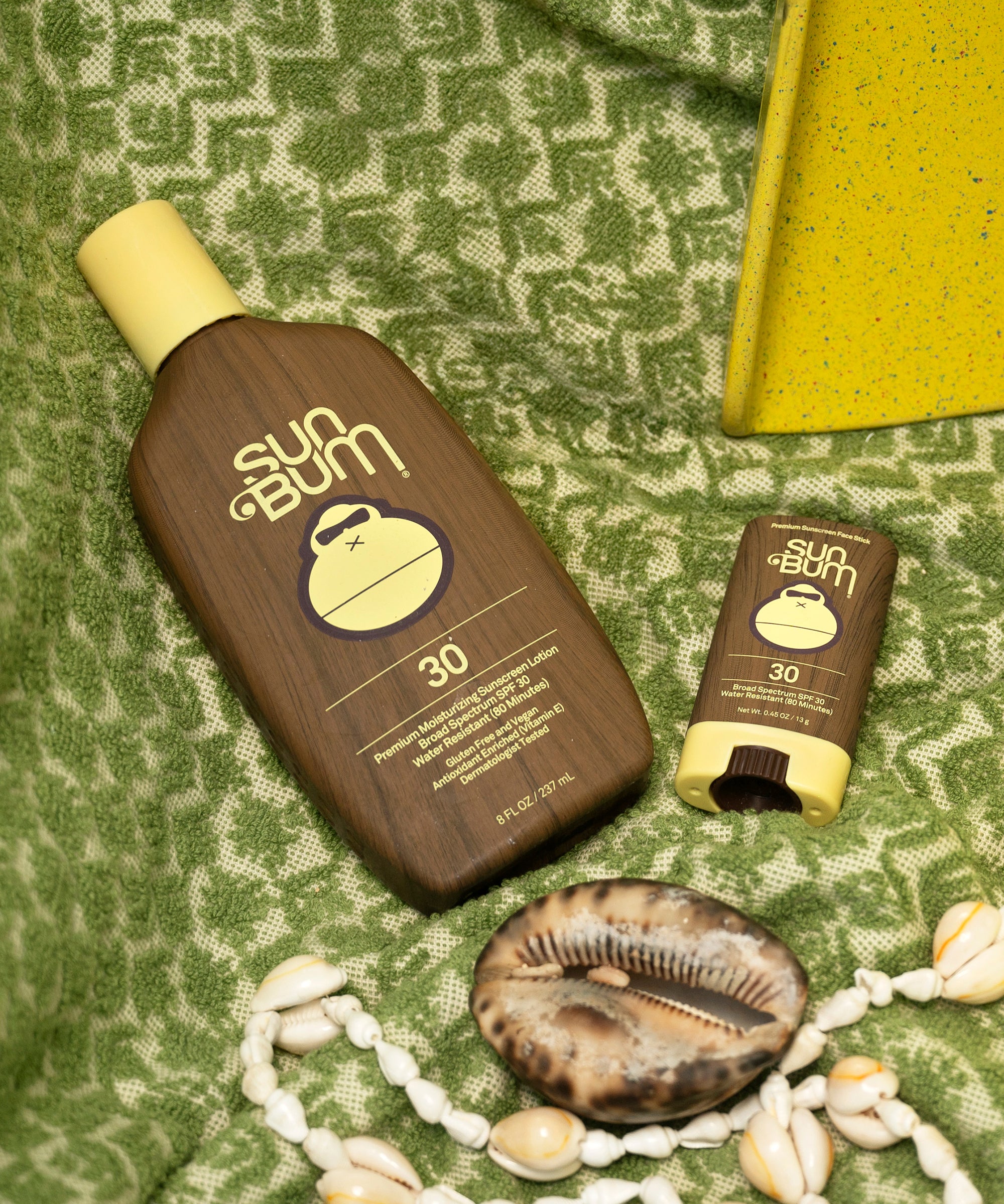 Sun Bum sunscreen bottle and stick on a green textured surface with shells and a yellow object.