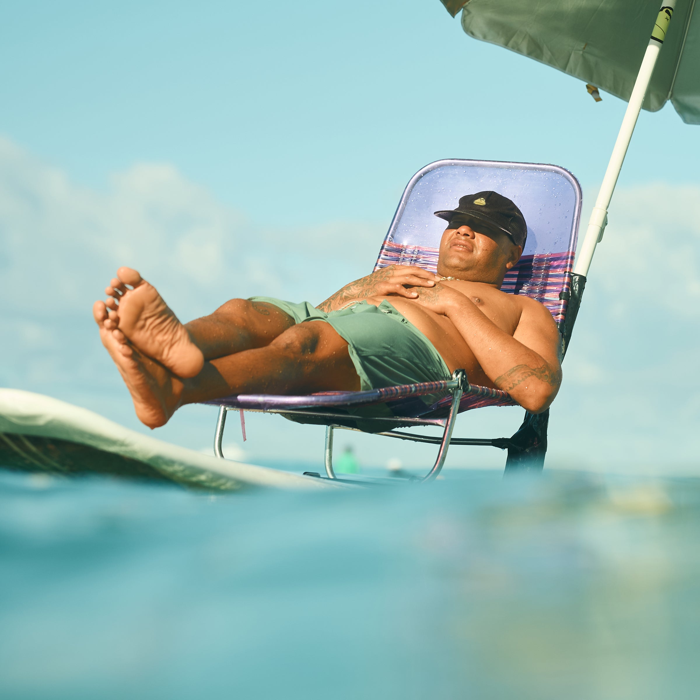 Man relaxing on a beach chair balanced on a surfboard in the ocean under an umbrella – Tanning Collection - Sun Bum