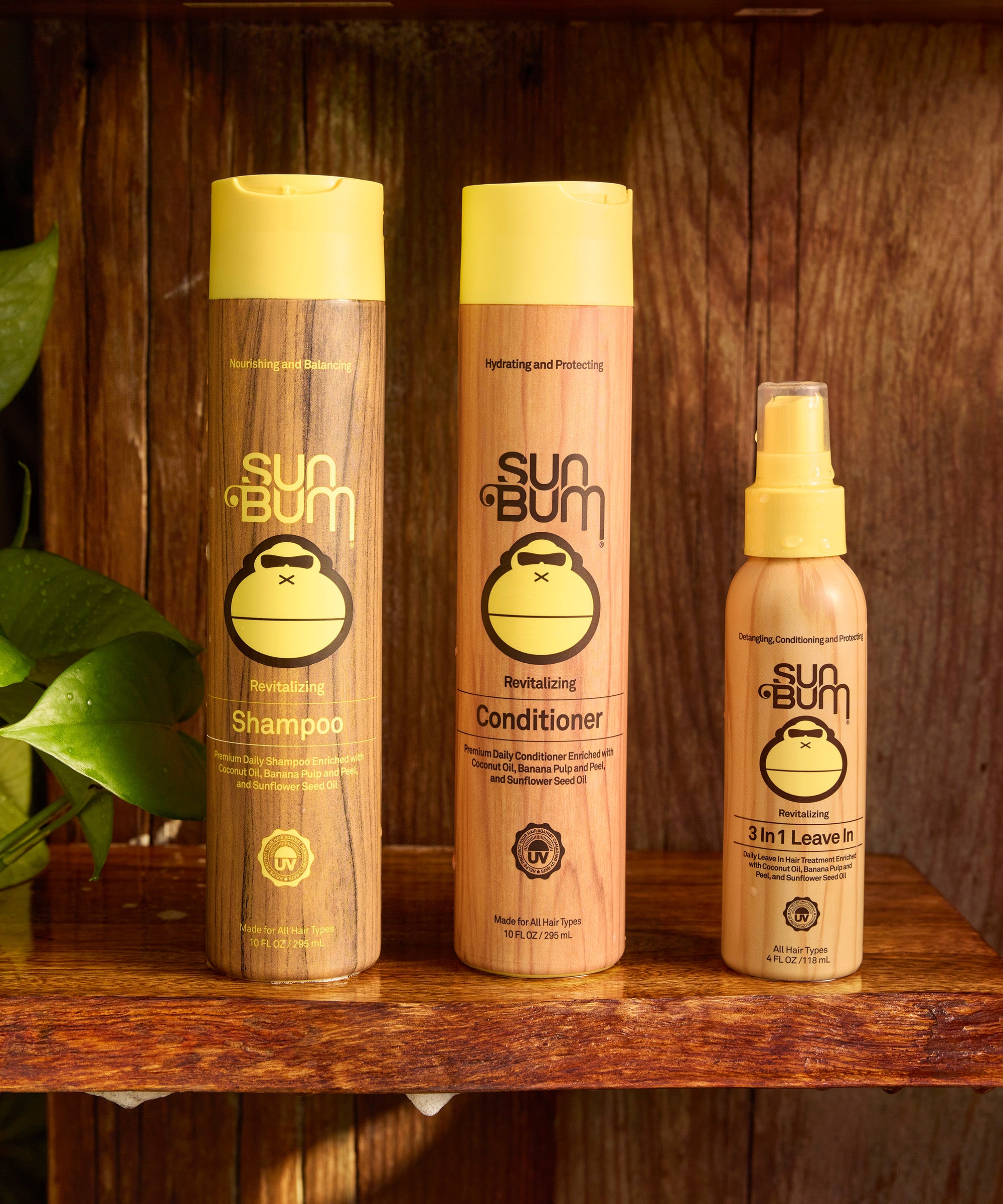 Revitalizing Shampoo bottle alongside matching conditioner and leave-in treatment, featuring brown and yellow packaging with monkey logo, displayed on wooden shelf with green plant in background - by Sun Bum