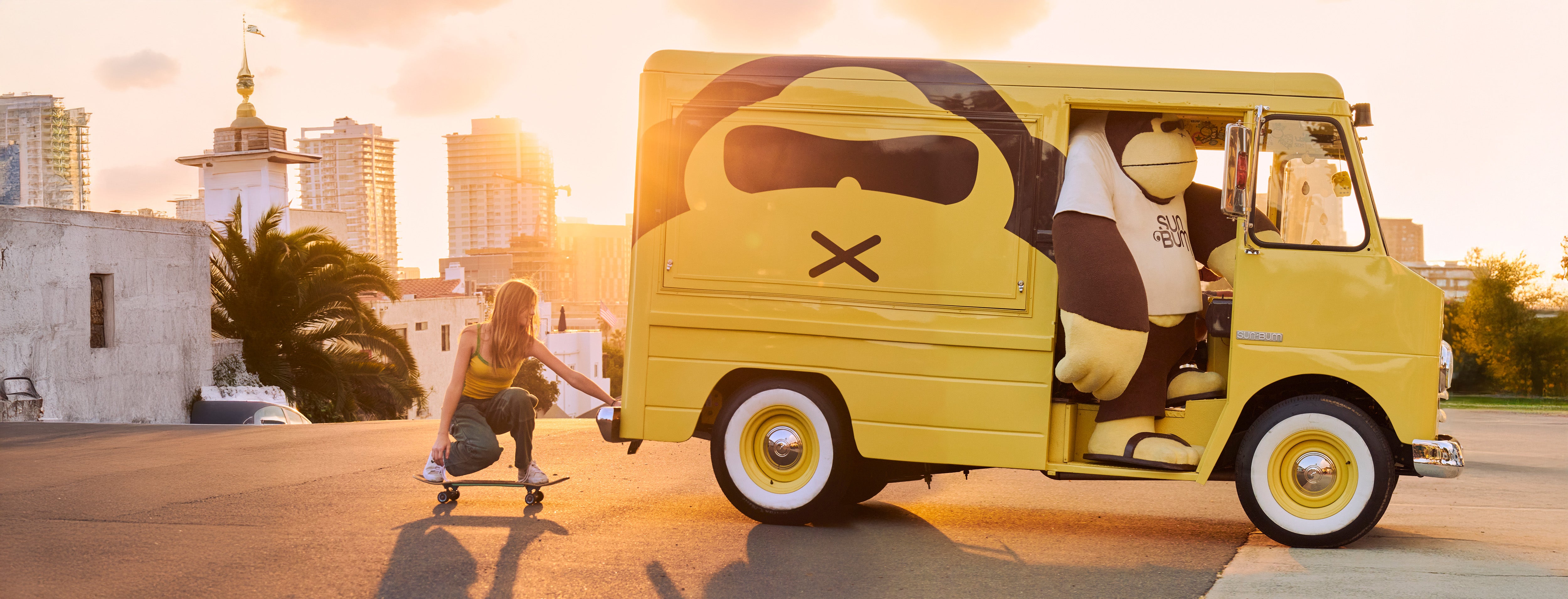 Girl skateboarding at sunset while holding onto a yellow Sun Bum van with mascot inside – Sun Bum.
