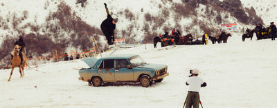 Person performing a snowboard trick over an old car in a snowy landscape with onlookers and a cameraman.