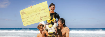 Three people on a beach holding a large check and a surfboard with a cartoon character.