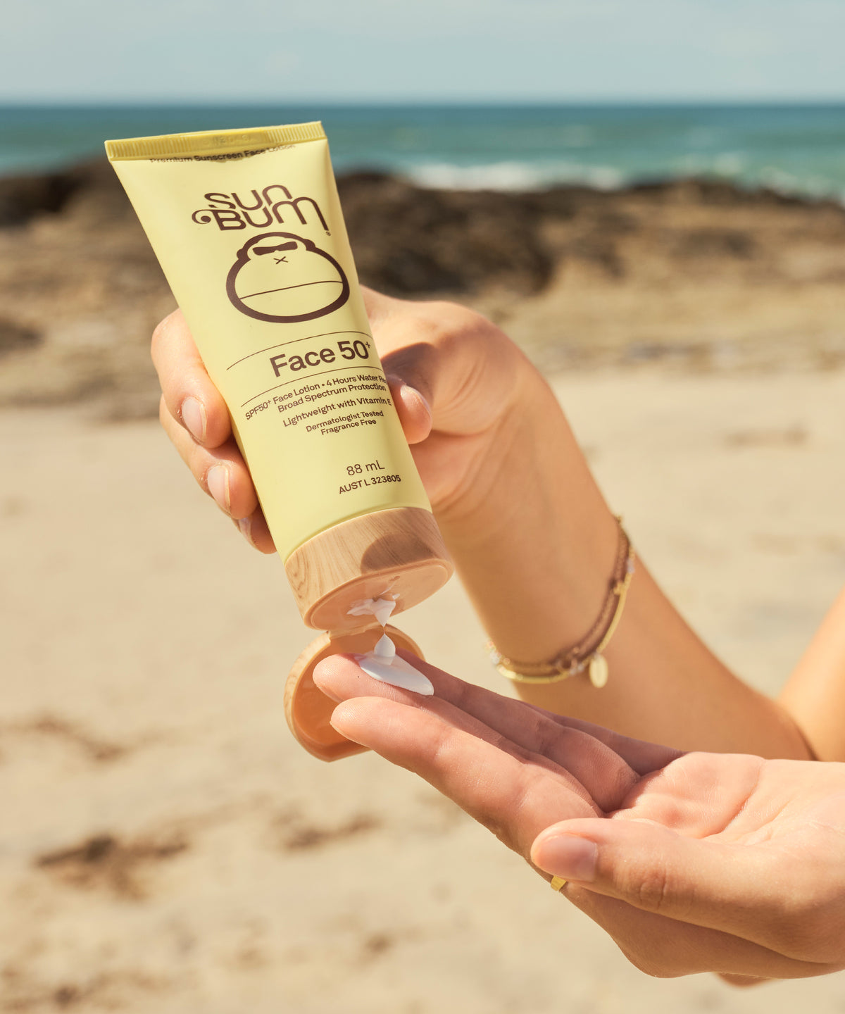 A person at a beach holds a tube of Sun Bum Face 50 sunscreen and dispenses some white lotion into their hand, with the ocean and sand blurred in the background.