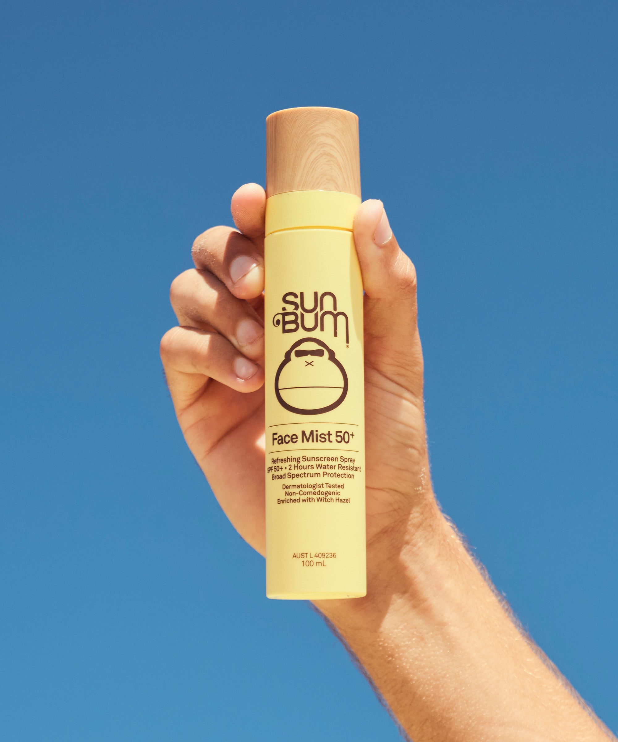 Hand holding a Sun Bum Face Mist 50+ spray bottle against a blue sky