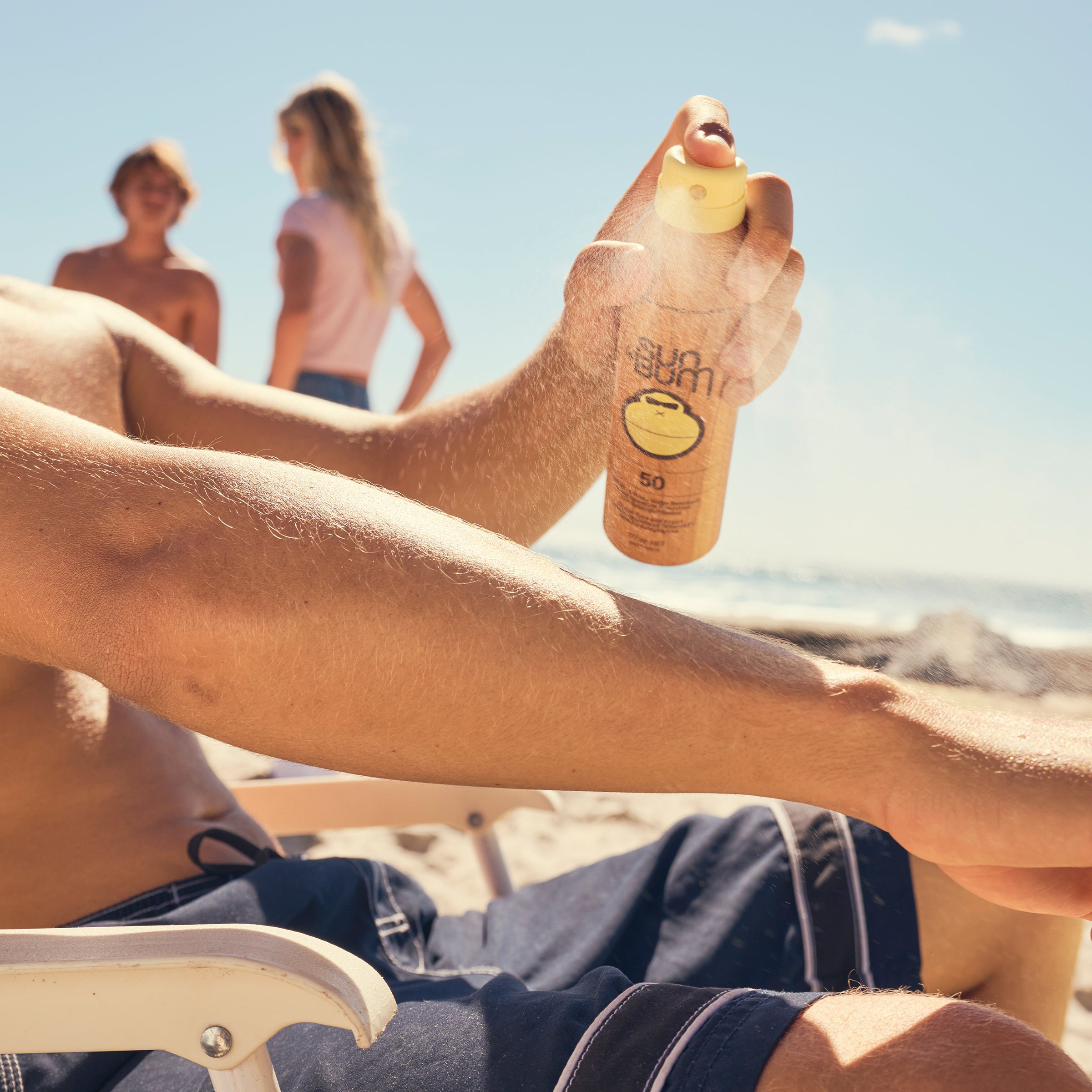 Person applying sunscreen spray on a beach with two people in the background.