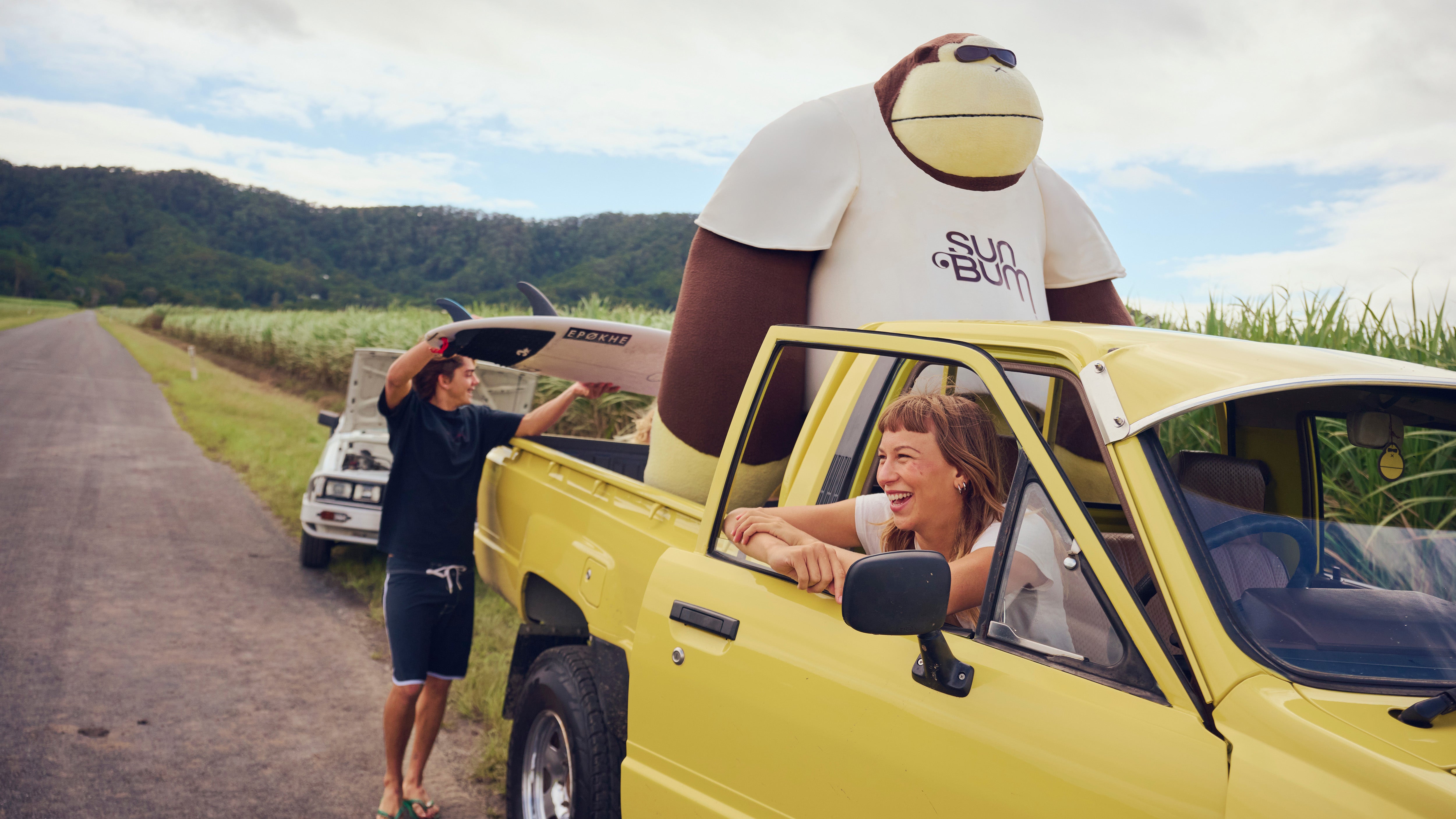 Two people in a yellow car with a large inflatable monkey in the back, on a road trip.