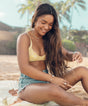 A young woman with long dark hair smiling and sitting on the beach. Ingredients like Sunflower Seed Oil, Jojoba Seed Oil, and Abyssinian Seed Oil in Sun Bum's Revitalizing Protect & Repair Oil help strengthen, hydrate, and protect hair.