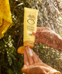 Yellow tube of Revitalizing Hair Mask held in sunlit hands with water droplets splashing in background, creating a refreshing summer scene with golden light filtering through leaves - by Sun Bum