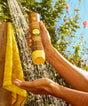 Hands holding a brown and yellow bottle of Revitalizing Shampoo against a sunny outdoor backdrop with tree and yellow towel, showcasing the moisturizing hair care product by Sun Bum