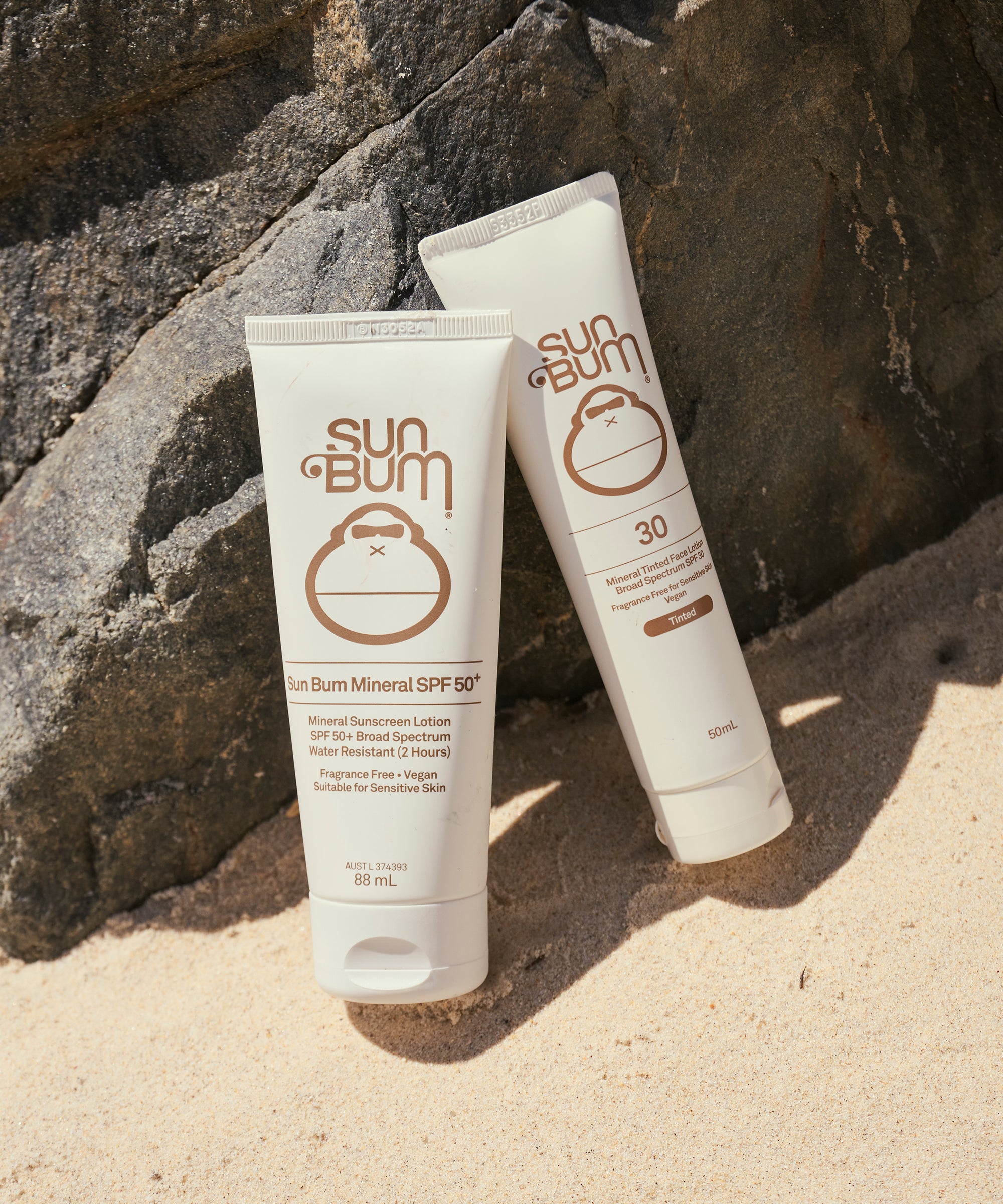 Two tubes of Sun Bum sunscreen on a sandy surface with rocks in the background