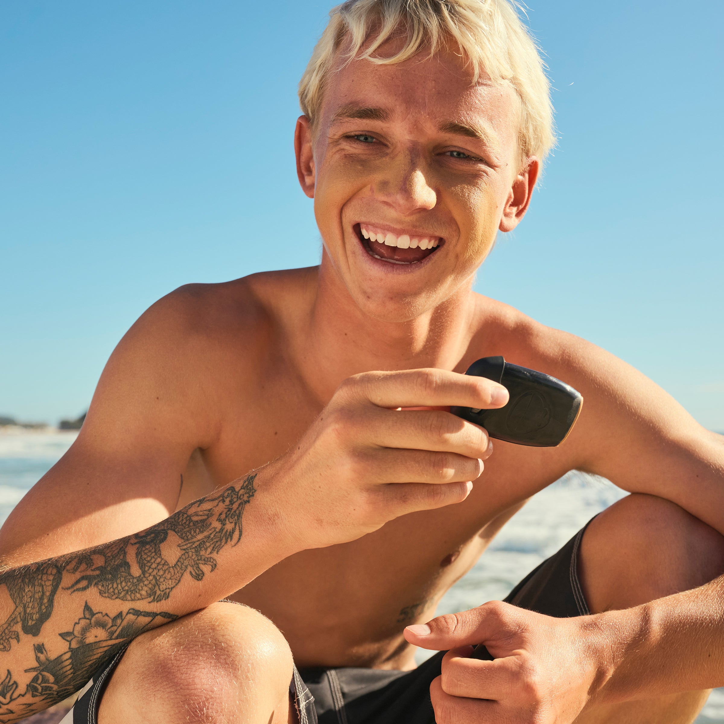 Man with tattoos on his arm sitting on a beach, holding a phone.