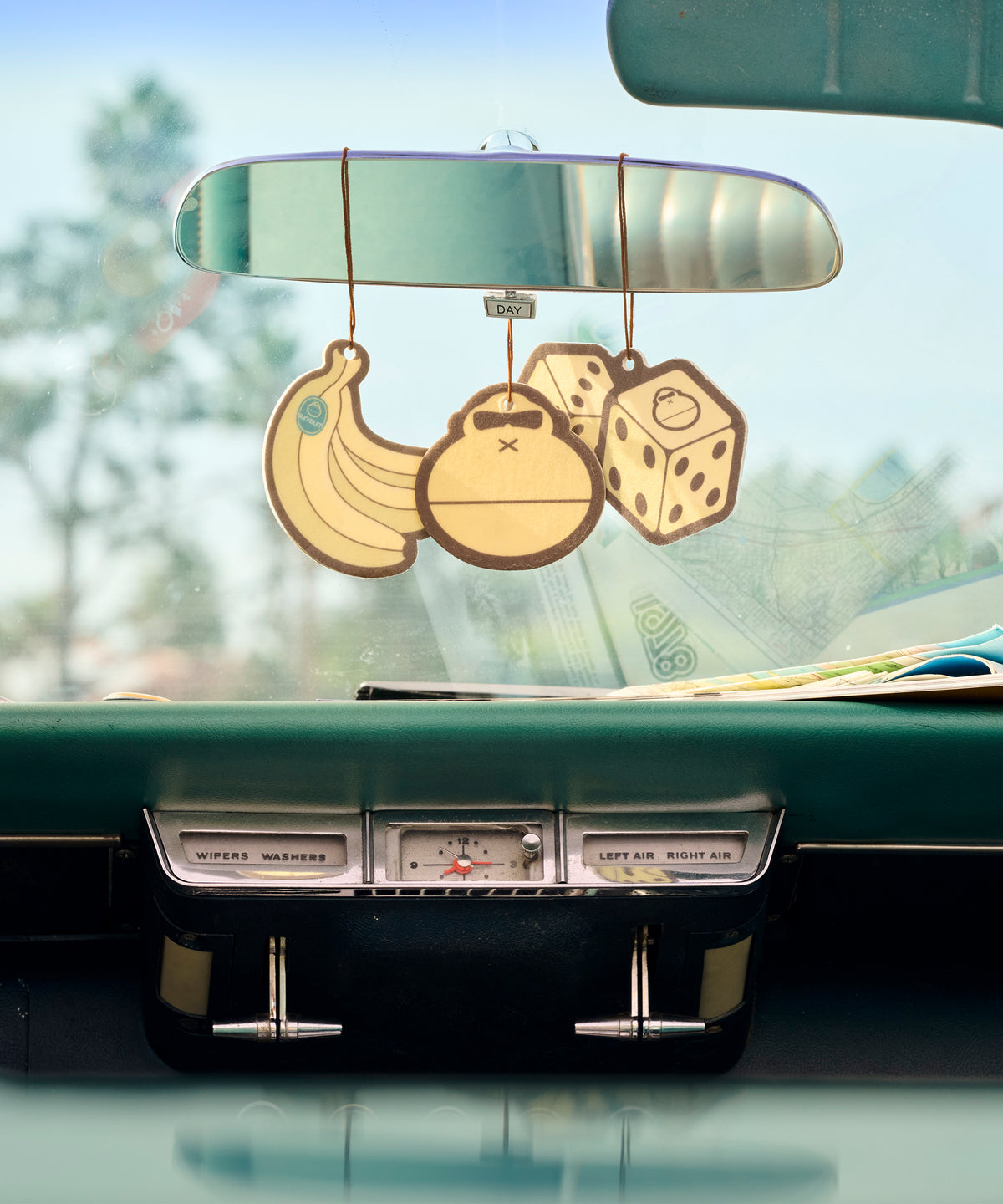 Three cartoon-style car air fresheners hanging from a rearview mirror with a blurred outdoor background.