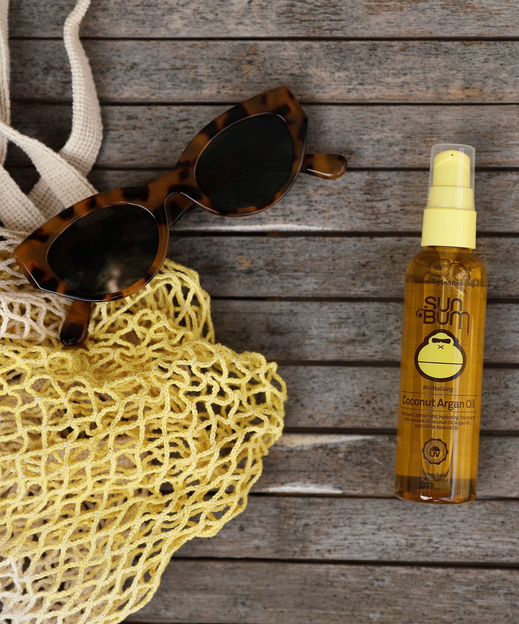 Coconut Argan Oil by Sun Bum placed on a wooden surface next to tortoiseshell sunglasses and a yellow mesh bag.