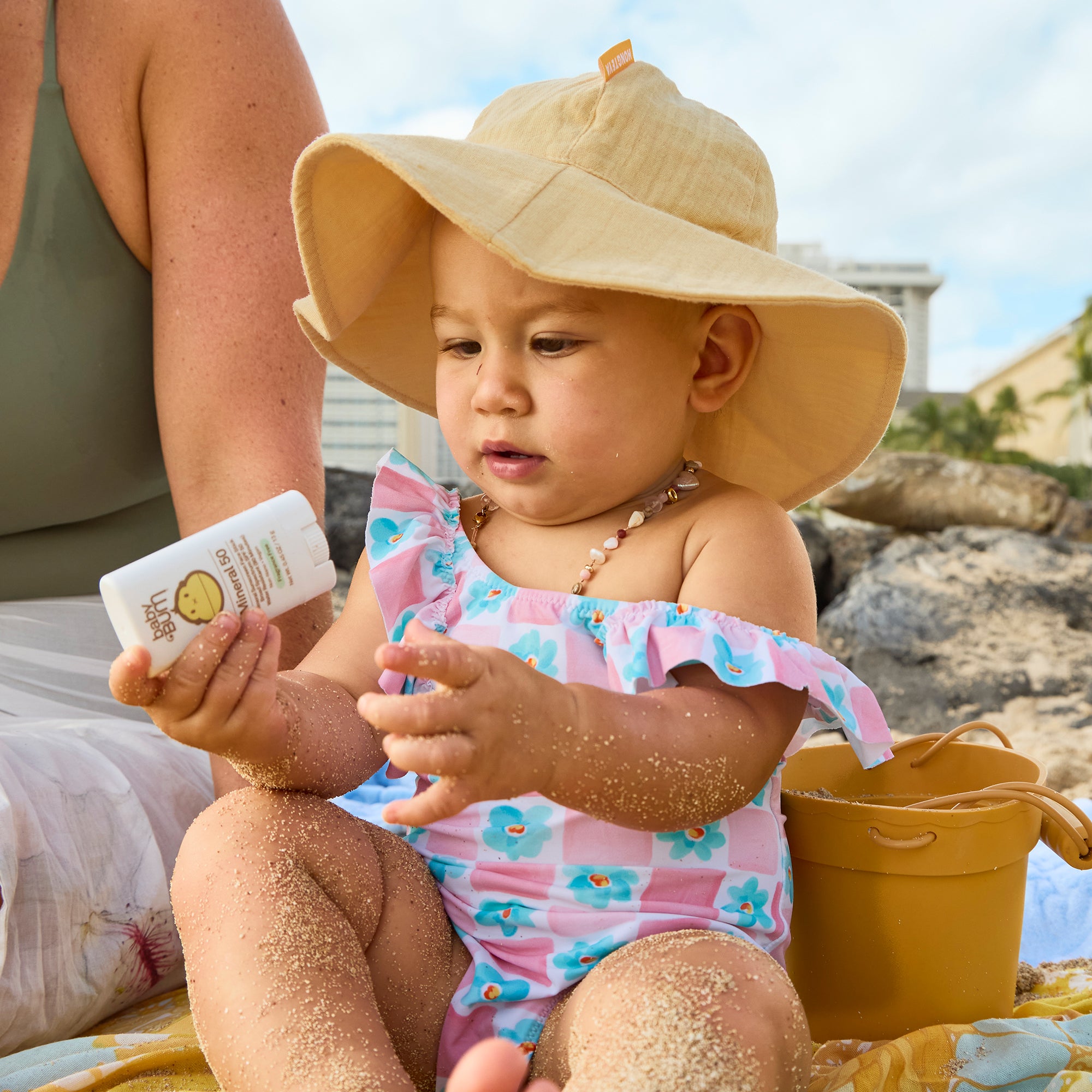 Baby in a sun hat and colorful swimsuit applying sunscreen on a beach.