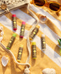 Colorful Original SPF 30 Sunscreen Lip Balm collection displayed on a yellow geometric patterned beach blanket with seashells and sunglasses, featuring coconut flavor and protective ingredients. Perfect for sunny day lip care by Sun Bum.
