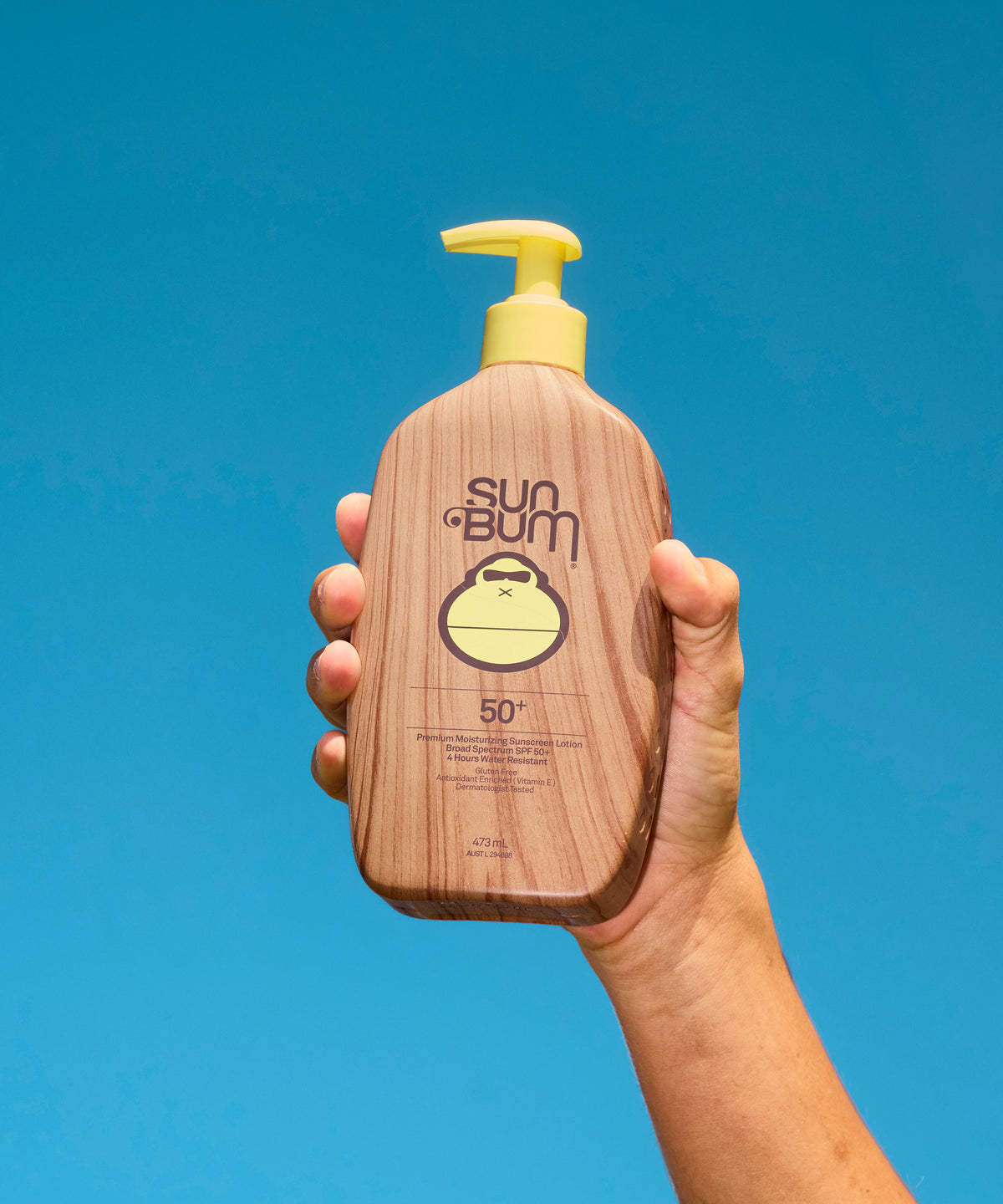 A hand holding a bottle of Sun Bum sunscreen against a solid blue background. The bottle has a brown wood-grain pattern with a yellow pump dispenser and a yellow gorilla logo.