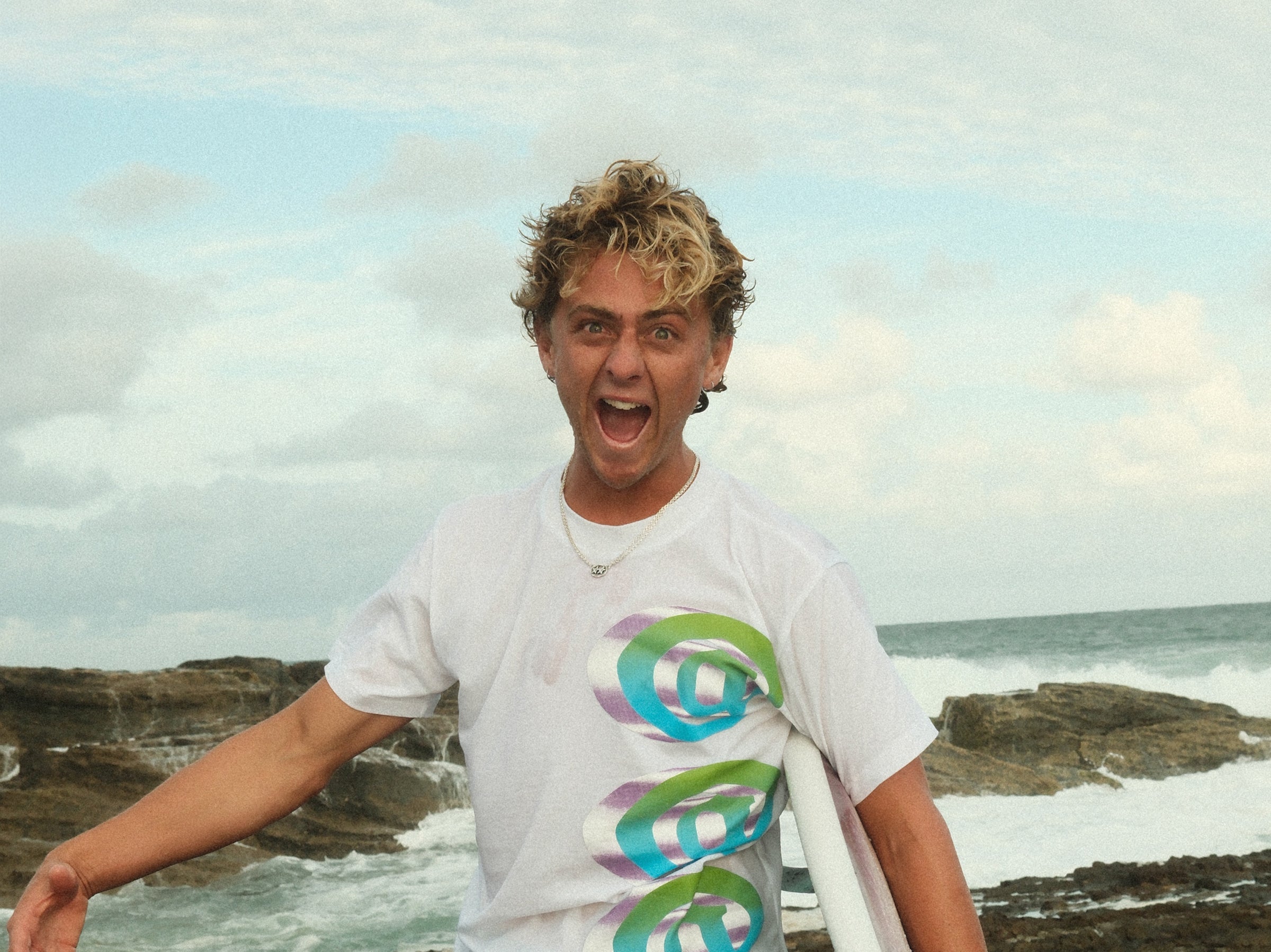 Person with a white t-shirt featuring colorful spiral design standing on a rocky beach.