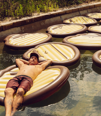 Person relaxing on a pool float surrounded by Sonny face inflatables in a backyard pool scene - Merch Collection - Sun Bum