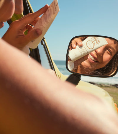 Person holding a bottle of sunscreen next to a car's side mirror with a reflection of the same bottle.