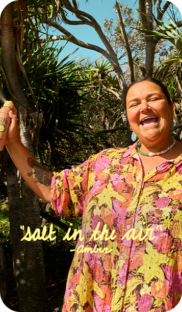 A woman outdoors wearing a colorful floral button-up shirt with pink, yellow, and orange tones, standing in front of trees and plants under a blue sky. Yellow text overlay reads 'salt in the air -Amber'.