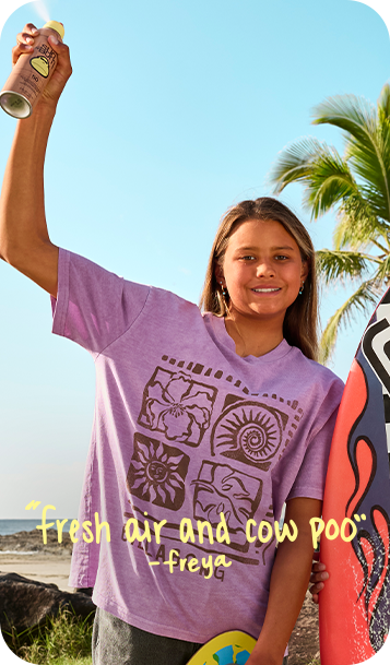A woman outdoors on a beach holding a surfboard and a spray can, wearing a lavender graphic T-shirt with a brown geometric and floral print. Yellow text at the bottom reads 'Fresh air and cow poo' and 'freya'.