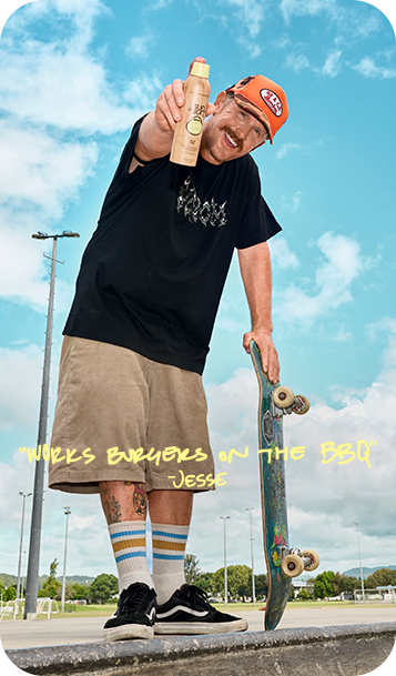 A man outdoors wearing a black graphic t-shirt, beige shorts, striped socks, and black sneakers, holding a skateboard and pointing a bottle toward the camera. The background shows a blue sky with clouds and a park setting.