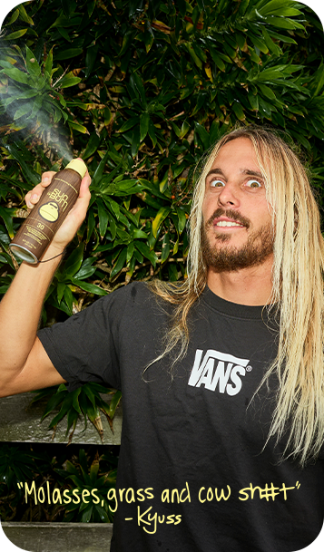 A man with long blonde hair and a beard holds up a spray can of Vans 'Molasses, grass and cow sh*t' against a background of green foliage. He is wearing a black Vans t-shirt with a white logo.