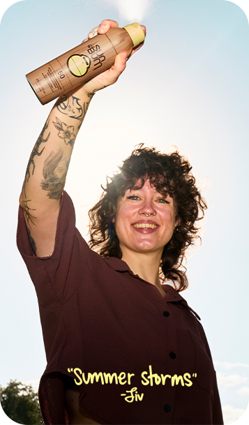 A smiling woman outdoors raises a brown spray bottle labeled 'Summer Storms' by Fin. She has curly dark hair, visible tattoos on her arm, and is wearing a dark brown button-up shirt against a bright sky background.