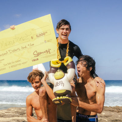 Three people on a beach with one holding a large check and another holding a toy, with ocean waves and sky in the background.