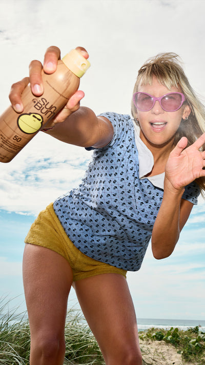 A woman outdoors against a cloudy blue sky holds up a tan spray bottle toward the camera. She is wearing pink sunglasses, a blue patterned top, and yellow shorts.