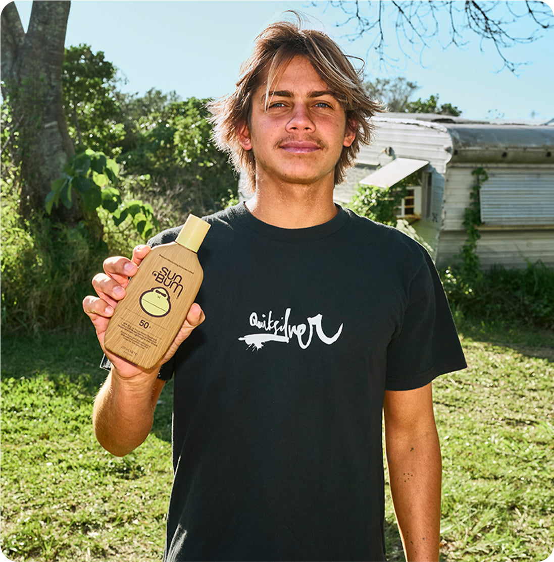 A man outdoors in a grassy area holds up a bottle of Quiksilver sunscreen. He is wearing a black Quiksilver t-shirt and standing in front of a rustic house and trees.