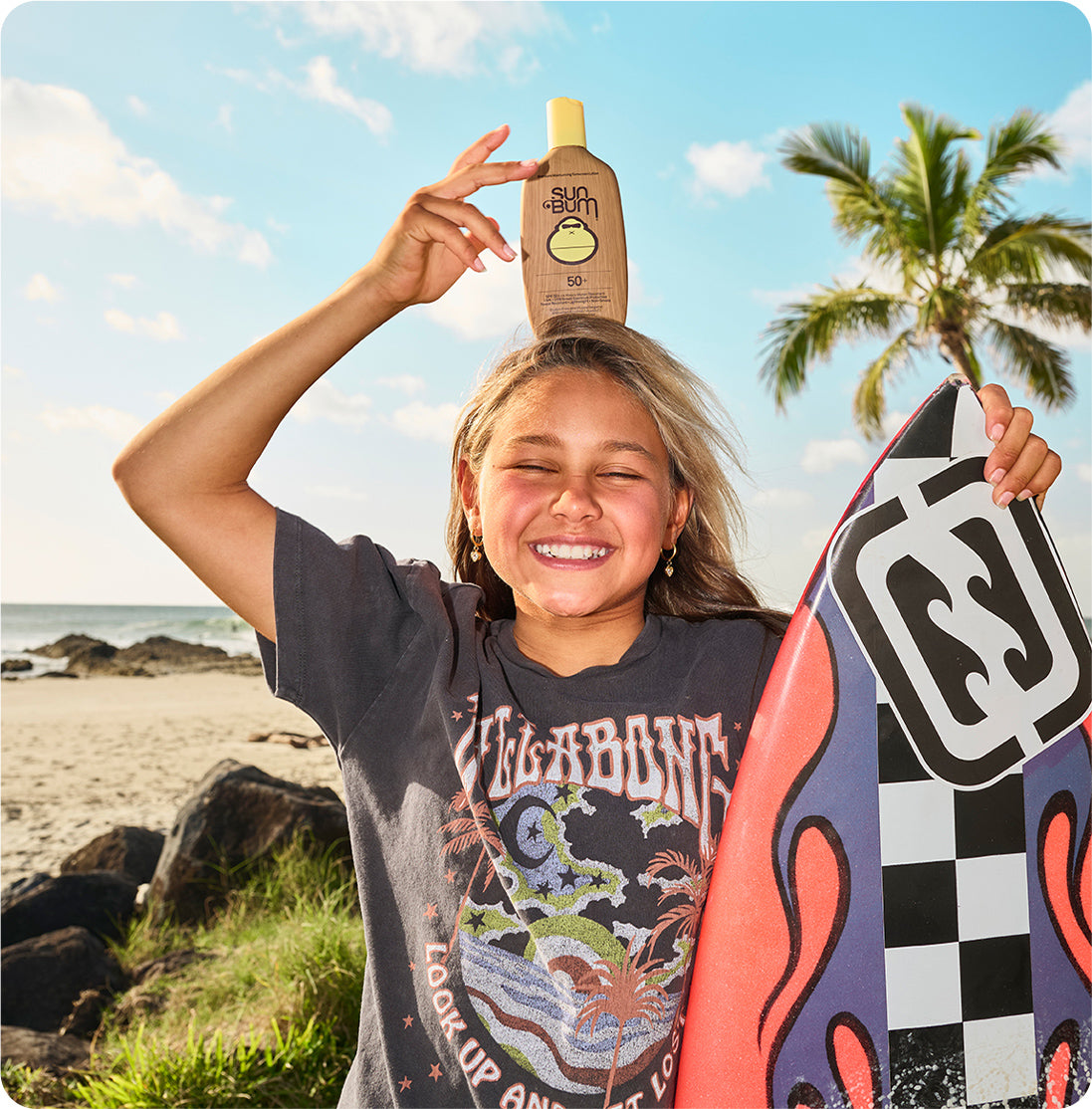 A smiling woman on a beach balances a bottle of Sun Bum sunscreen on her head while holding a colorful surfboard. The background shows the ocean, sand, rocks, and a palm tree under a blue sky.