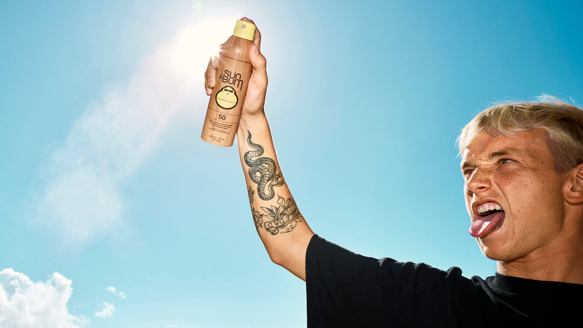 A man with blonde hair and arm tattoos holds up a bottle of sunscreen against a bright blue sky with the sun shining. The bottle is tan with a gold cap and a circular logo that appears to read 'Sun'.