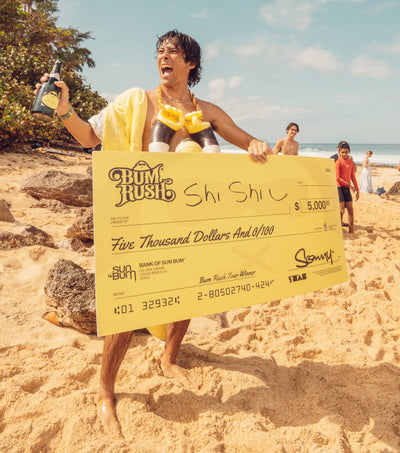 Surfer celebrates on the beach with a giant check and champagne after winning the Bum Rush Tour – Discover Collection - Sun Bum.