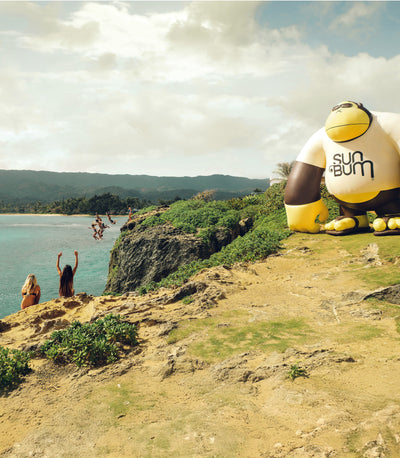 Friends cliff jumping by the ocean next to giant inflatable Sun Bum gorilla – Discover Collection - Sun Bum.