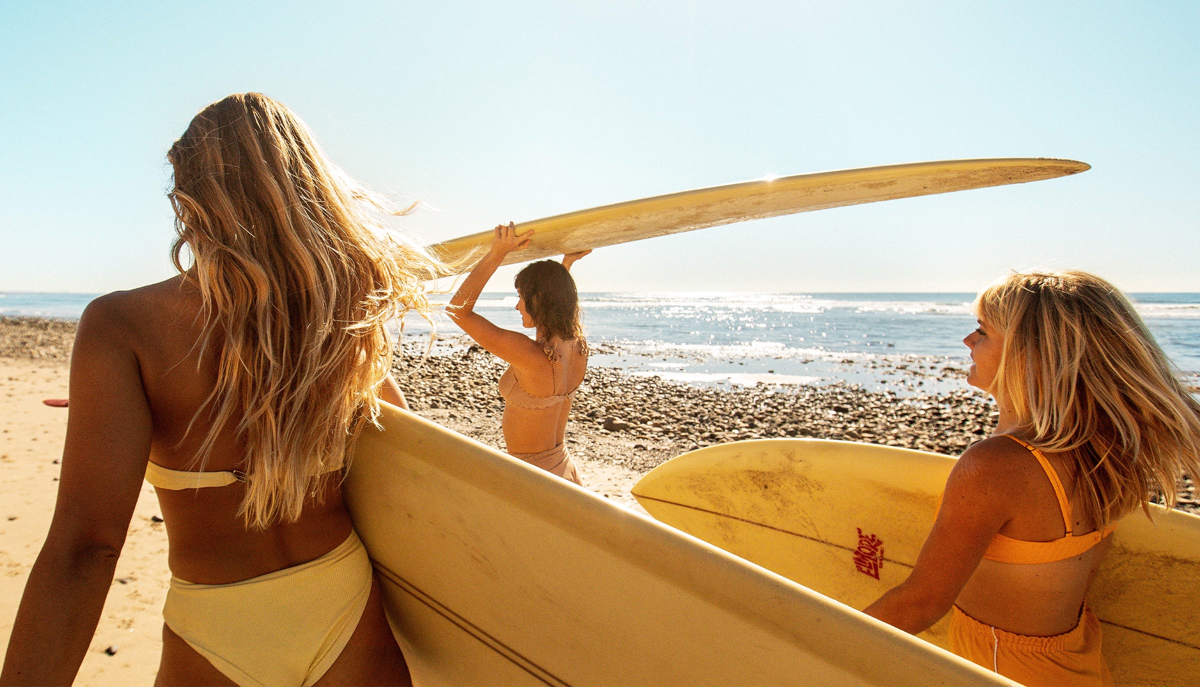 Women carrying surfboards toward the ocean under the sun, showing off healthy beach hair – Hair Collection - Sun Bum.