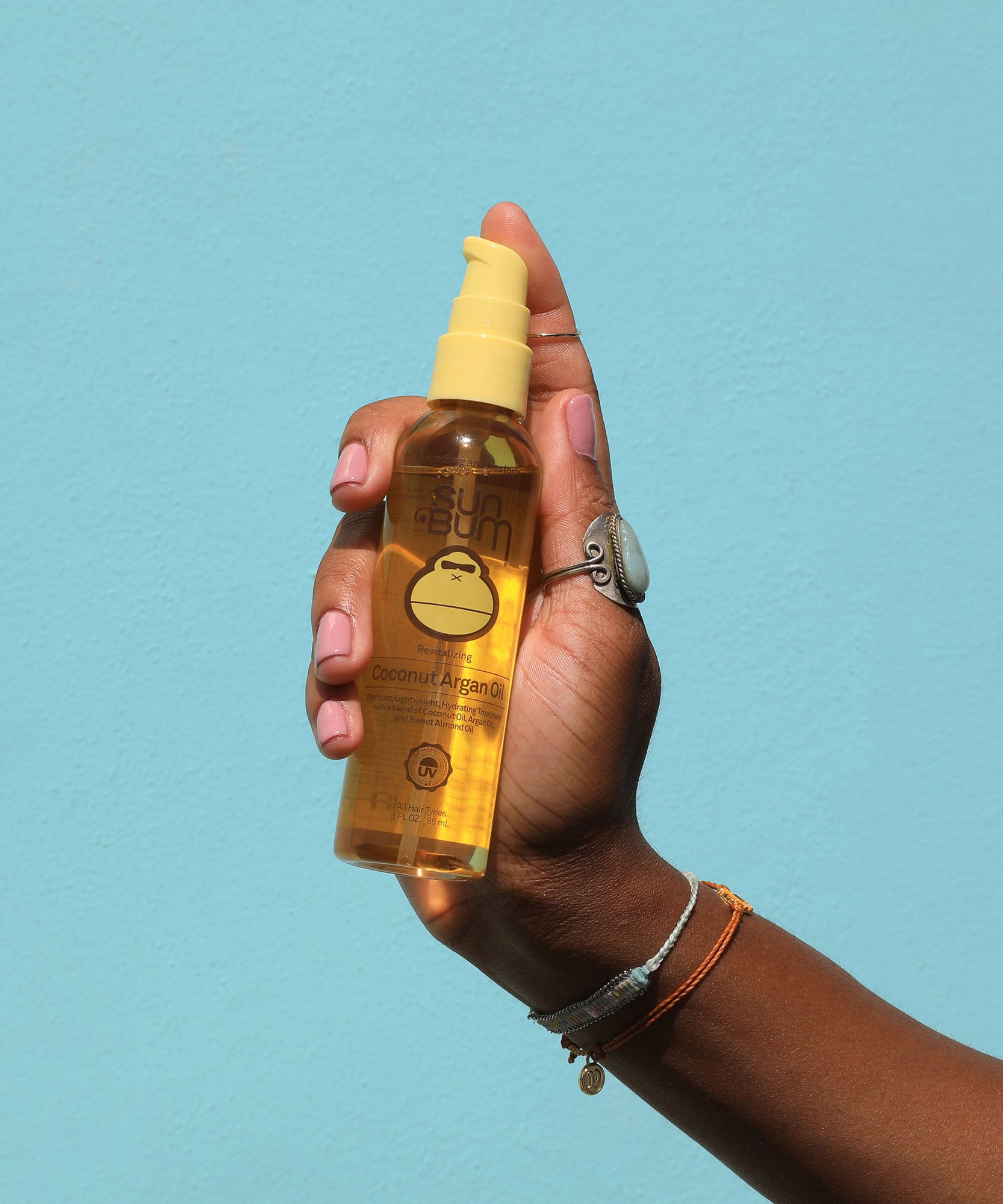 Hand holding a yellow bottle of Coconut Argan Oil against a light blue background, Sun Bum.