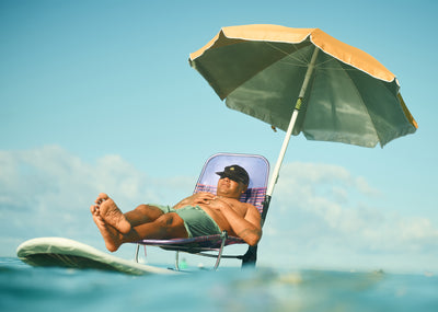 Man relaxing on a beach chair and umbrella balanced on a surfboard, showing creative sun protection from Know Your Spots Collection - Sun Bum.