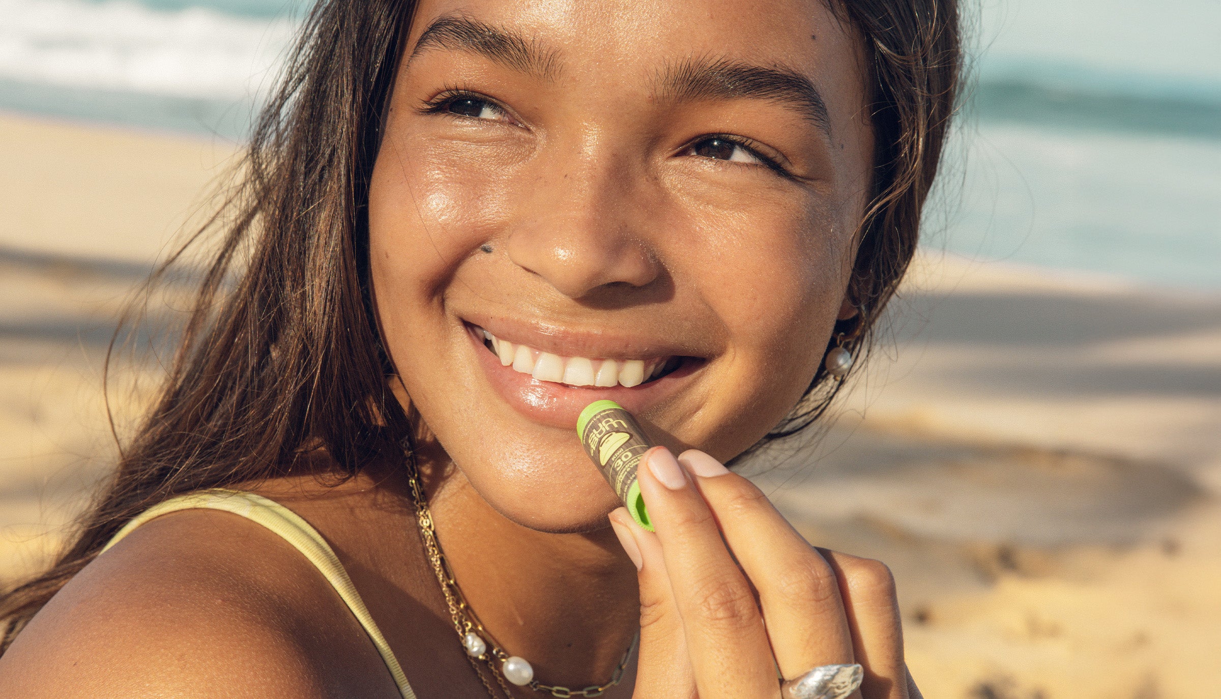Smiling woman applies SPF 30 lip balm at the beach for sun-safe, hydrated lips – OG Lip Balm Collection - Sun Bum.