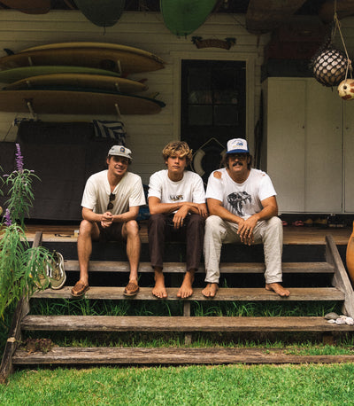 Three men sitting on wooden steps with surfboards in the background