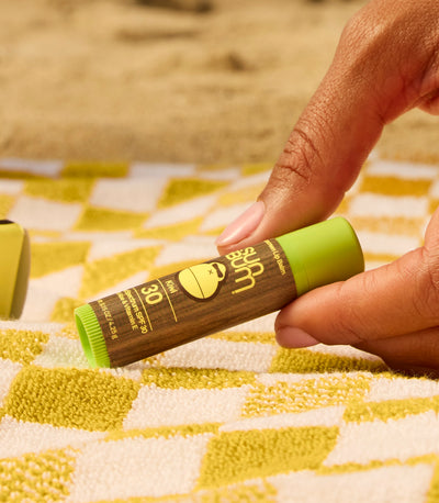 Close-up of hand picking up SPF 30 Kiwi lip balm on a beach towel, from the SB OG Lip Care Collection – Sun Bum.