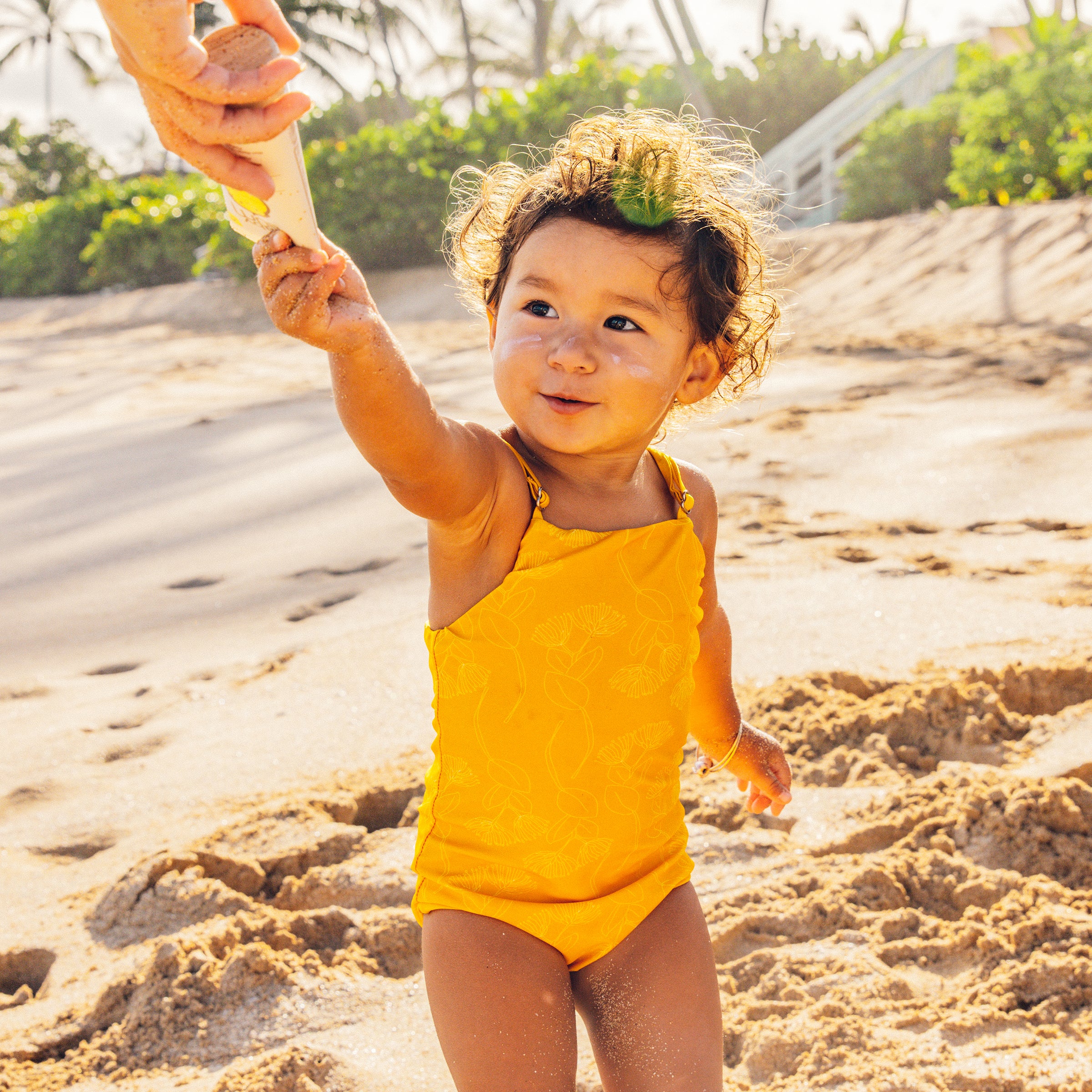 Smiling toddler with sunscreen on cheeks reaching for tube at the beach – Baby Bum Collection - Sun Bum.