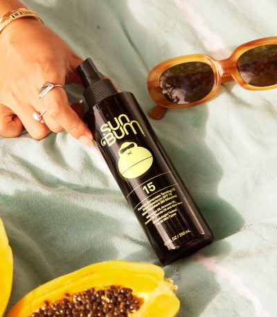 Sun Bum SPF 15 Tanning Oil on a beach towel with papaya and sunglasses, from the SB Tanning Oils and Lotions Collection – Sun Bum.