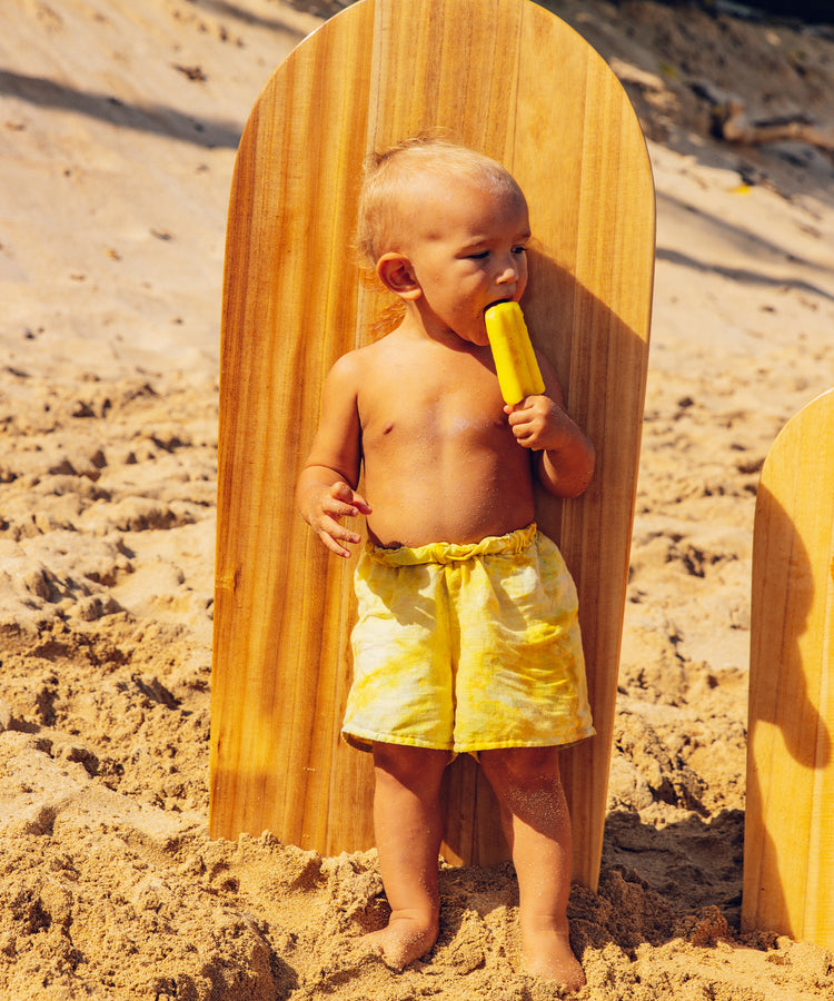 Toddler in yellow shorts enjoys a popsicle on the beach, leaning on a mini surfboard in the sand - Baby Bum Collection - Sun Bum
