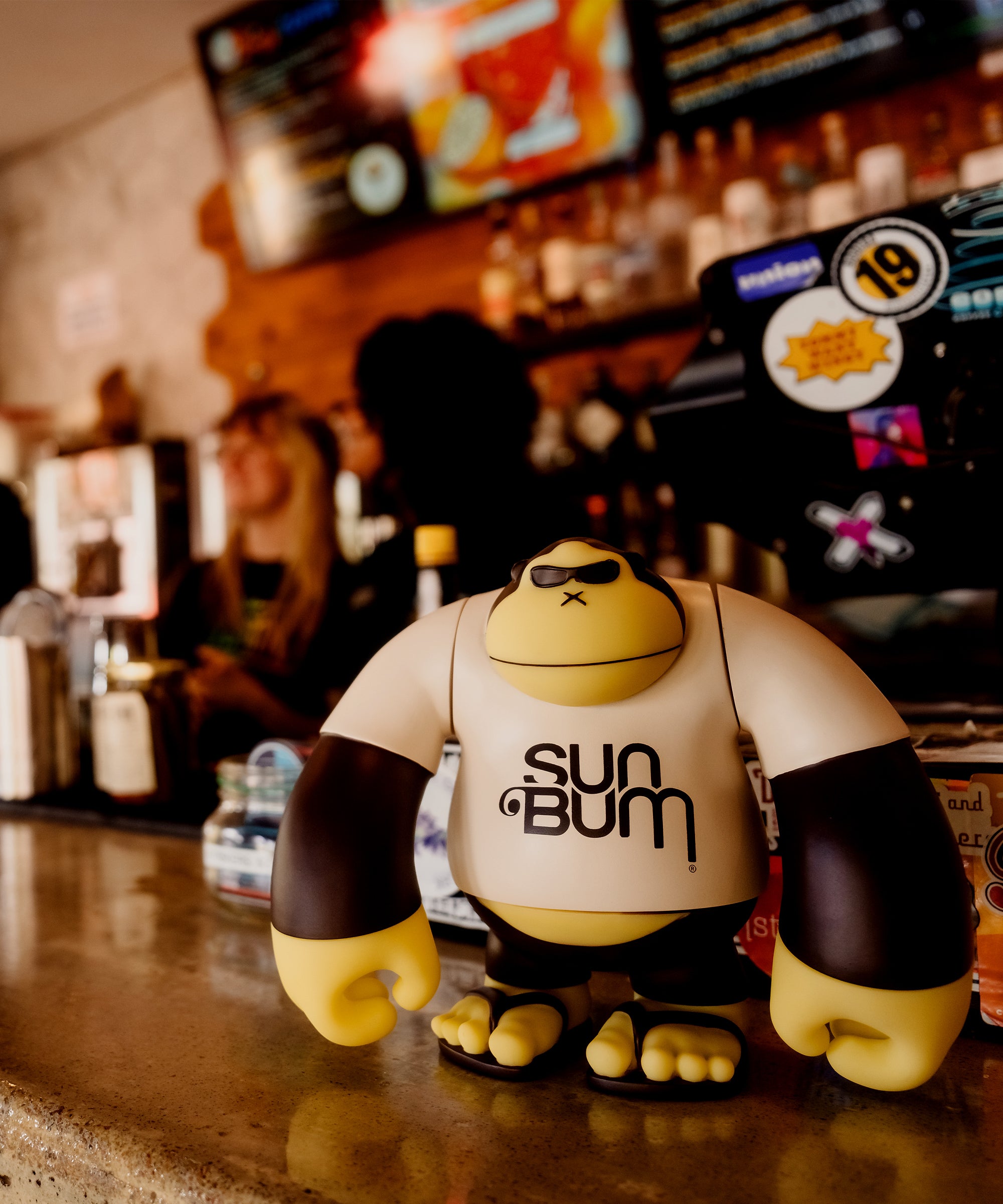Sun Bum mascot figure on a bar counter with bottles and staff blurred in background - Brand Vibes Collection - Sun Bum
