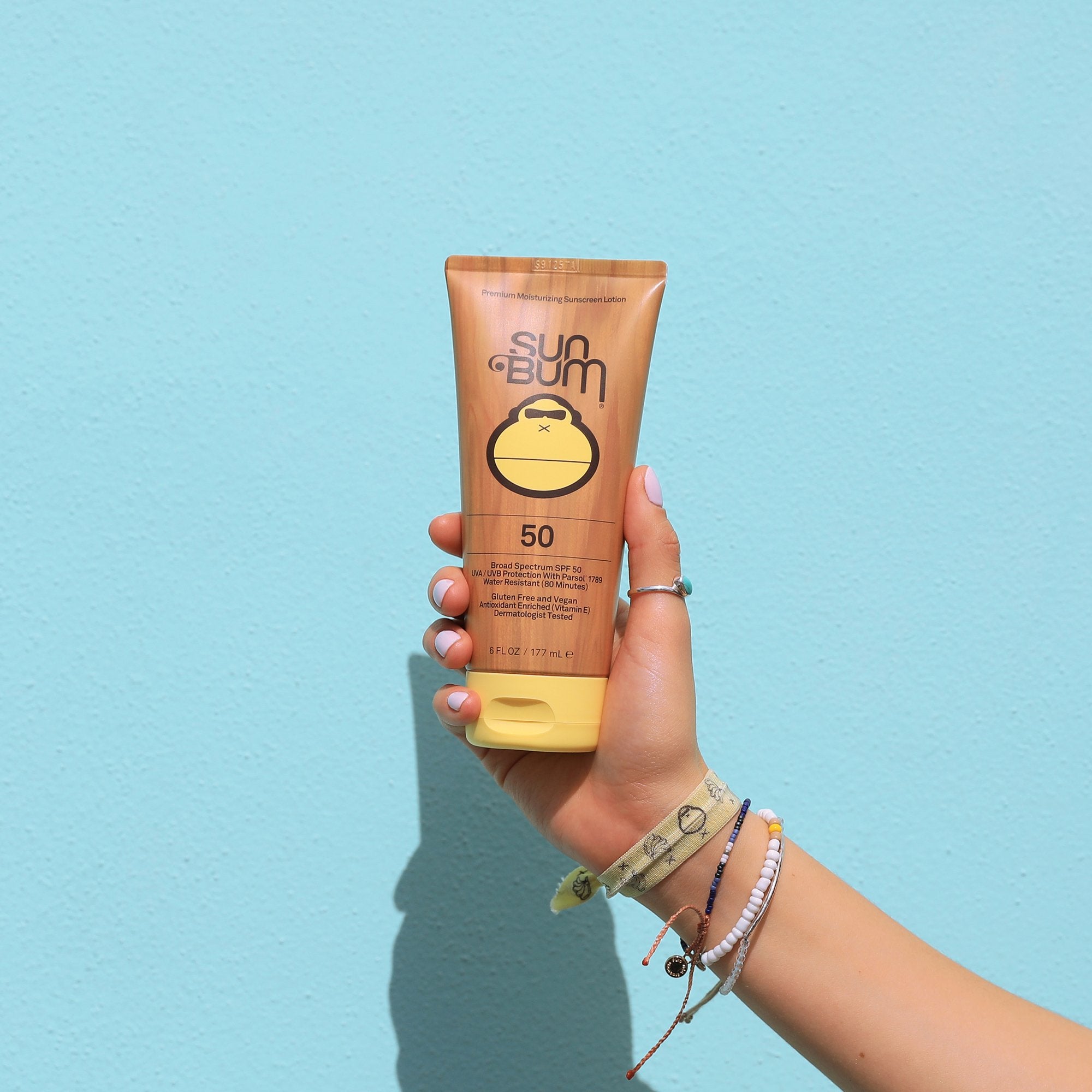 Hand holding Sun Bum SPF 50 sunscreen lotion against a blue wall, perfect for daily sun protection - Original Collection - Sun Bum